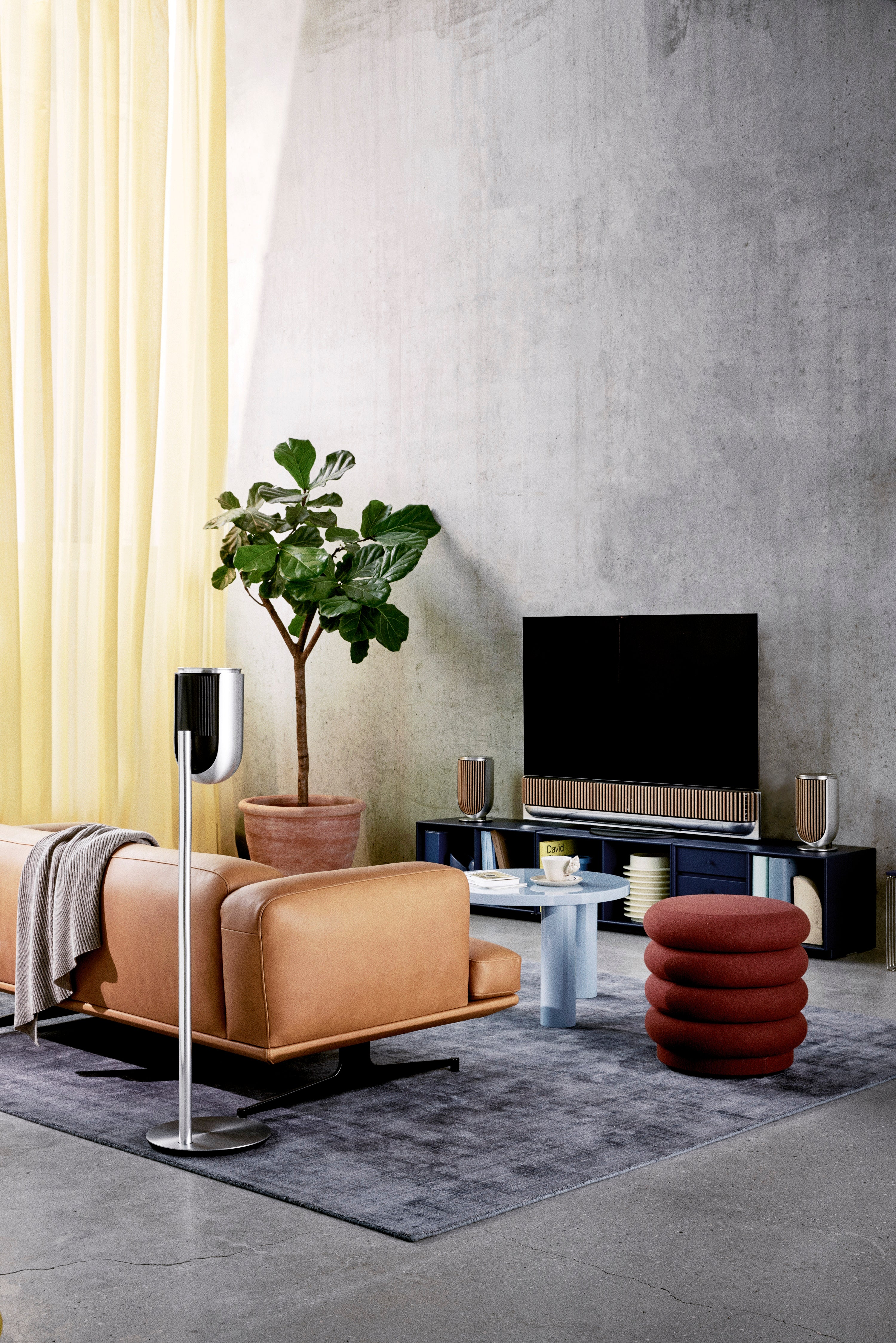Modernes Heimkino-Setup in einem Loft mit Sichtbetonwänden. Im Zentrum steht die Beovision Theater mit Eichenholz-Lamellen, flankiert von zwei Beolab 8 auf Tischstandfüßen. Im Vordergrund links markiert ein dritter Beolab 8 auf einem hohen Bodenstandfuß (Natural Aluminium/Black Fabric) die Rear-Position. Das Bild verdeutlicht die Flexibilität des Beolab 8 innerhalb eines konsistenten Design-Ökosystems.
