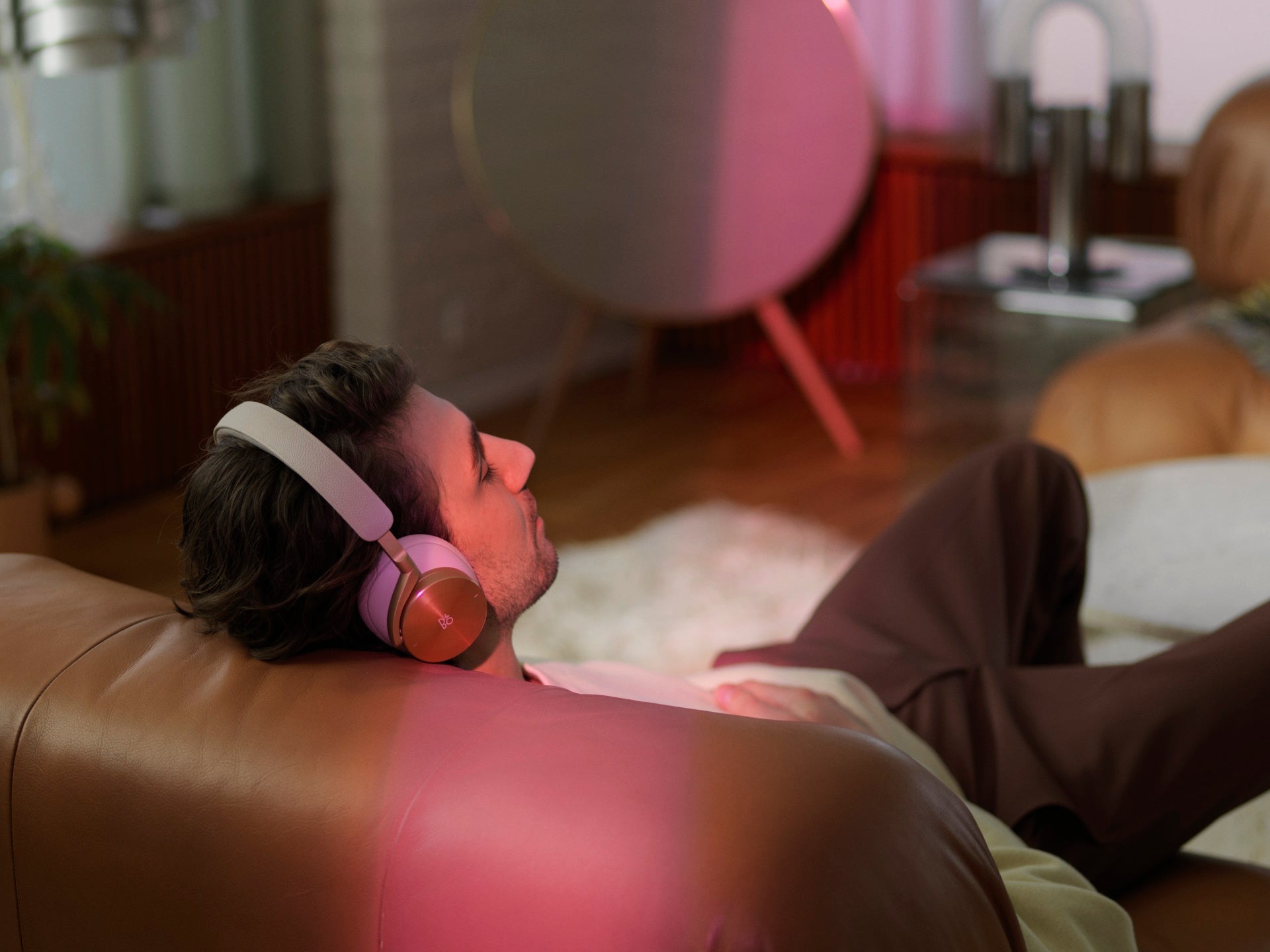 Lifestyle-Aufnahme eines Mannes, der entspannt auf einem braunen Ledersofa liegt und goldene Bang & Olufsen Over-Ear-Kopfhörer trägt. Im Hintergrund ist ein Beosound A9 Design-Lautsprecher in einem modern eingerichteten Wohnzimmer mit warmer, farbiger Akzentbeleuchtung zu sehen.