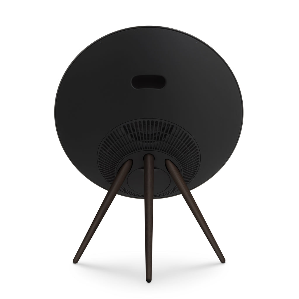 Zentrale Rückansicht der Bang & Olufsen Beosound A9 in Black Anthracite. Das Bild zeigt das nahtlose, schwarze Rückgehäuse mit dem ergonomischen Tragegriff, die kreisförmig angeordneten Belüftungsschlitze und die stabilen Stativbeine aus dunklem Nussbaumholz. Professionelles Produktfoto, freigestellt auf weißem Hintergrund.