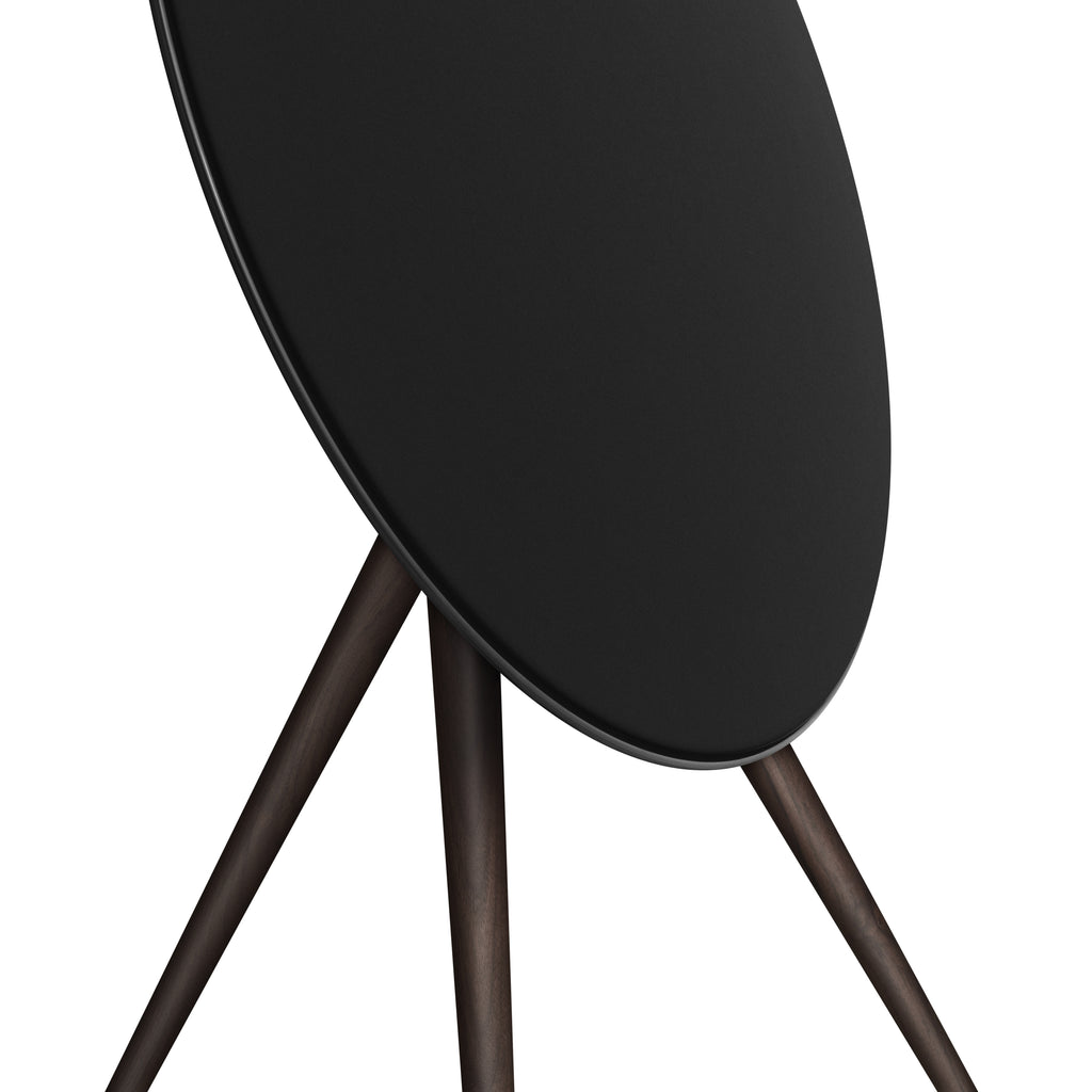 Detaillierte Nahaufnahme der Bang & Olufsen Beosound A9 in Black Anthracite. Der Fokus liegt auf dem Übergang zwischen dem anthrazitfarbenen Aluminiumring, der fein gewebten schwarzen Textilfront und den Beinen aus massiv dunklem Nussbaumholz. Die Aufnahme betont die edle Haptik und die erstklassige Materialverarbeitung.