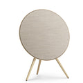 Frontansicht der Beosound A9 in Gold Tone. Das kreisrunde Design mit sandfarbenem Kvadrat-Bezug und hellen Eichenholzbeinen wirkt wie ein zeitloses Kunstobjekt.