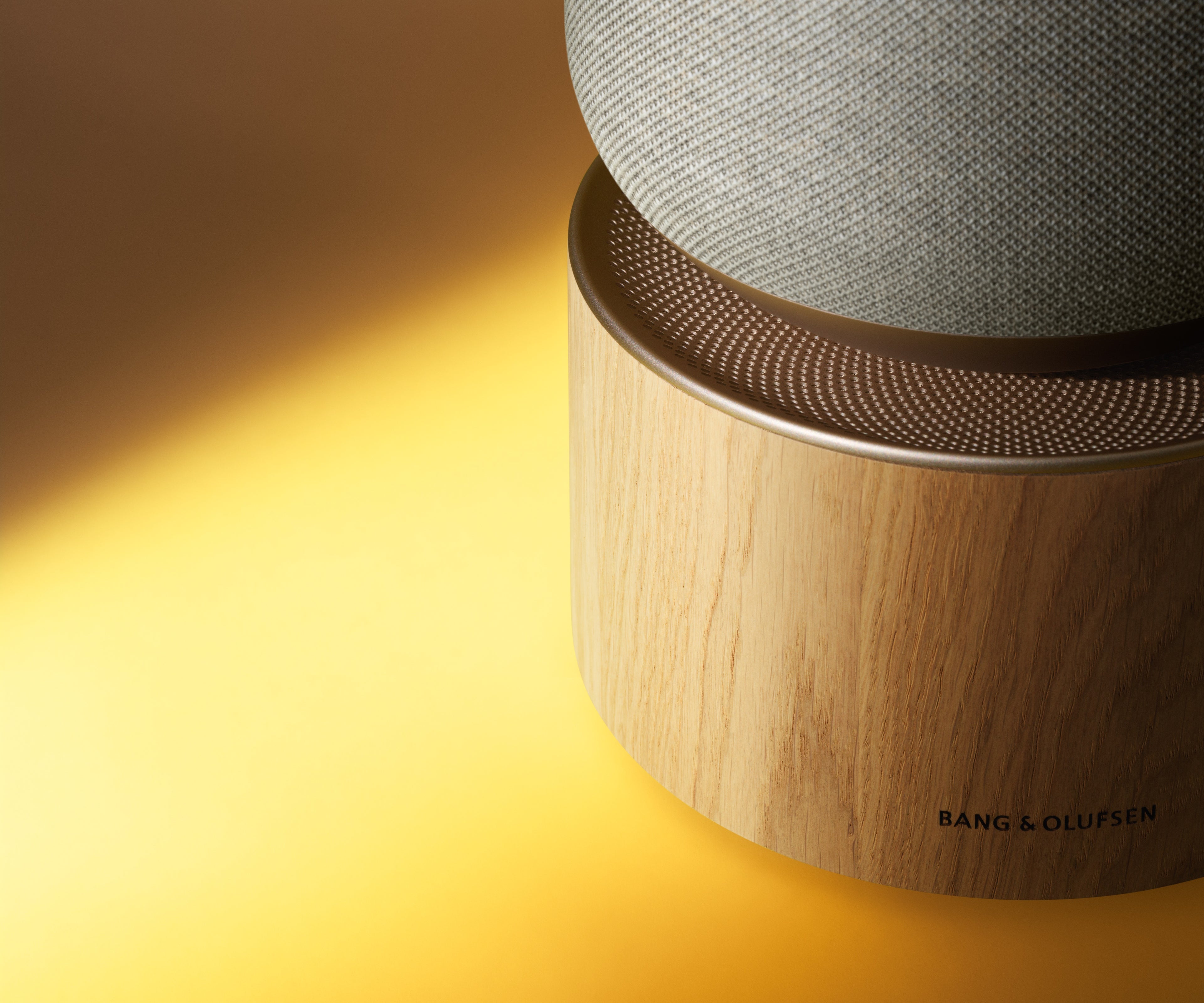 Nahaufnahme des Sockels eines Bang & Olufsen Beosound Balance Lautsprechers. Der Fokus liegt auf dem Sockel aus hellem Eichenholz mit dezent eingraviertem B&O-Logo, dem fein gelochten Aluminiumring in Gold-Tone und dem unteren Teil der beigen Stricktextil-Bespannung vor einem warmen, goldenen Lichtverlauf.