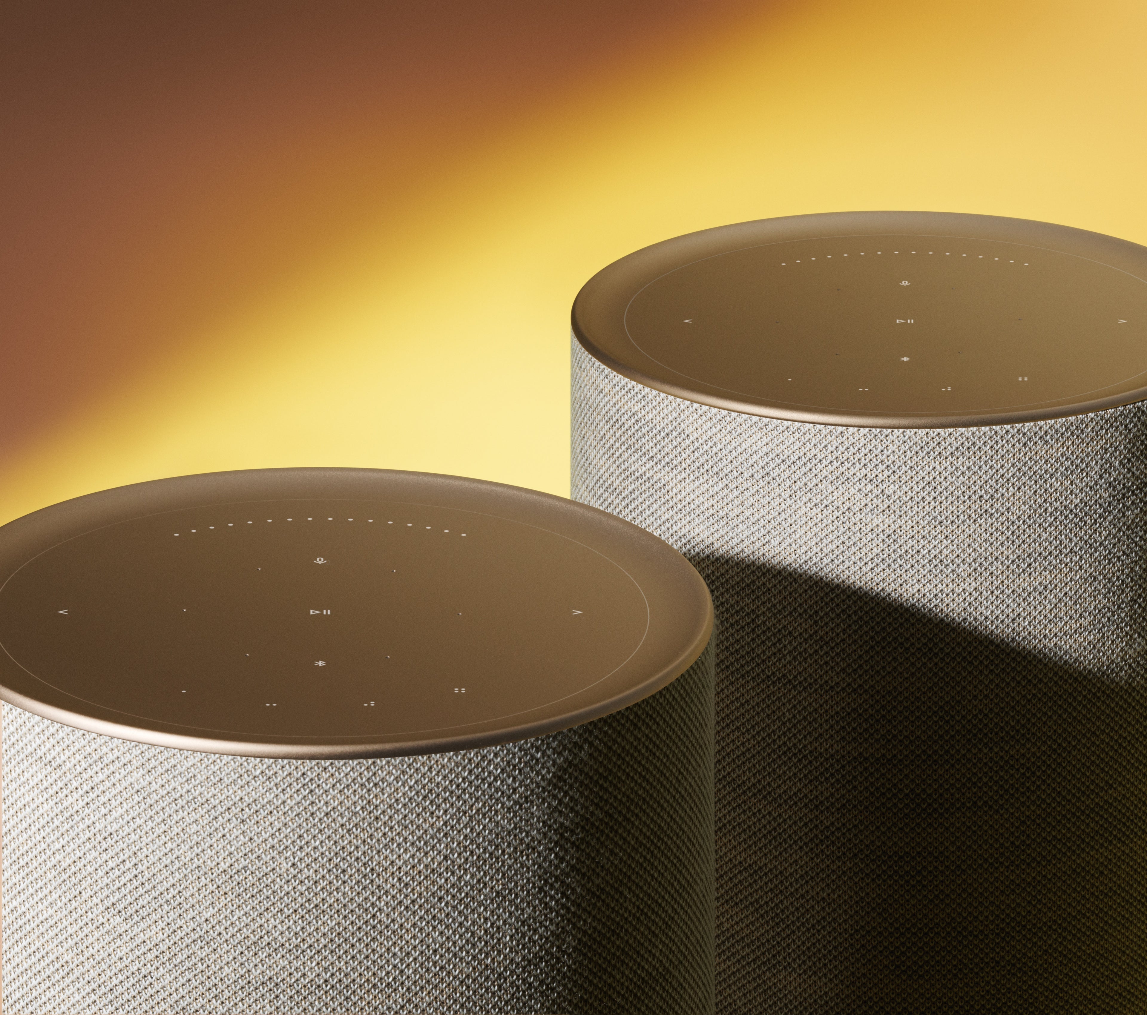 Nahaufnahme von zwei Bang & Olufsen Beosound Balance Lautsprechern als Stereo-Paar in Gold Tone. Der Fokus liegt auf der berührungsempfindlichen Oberseite aus mattem Aluminium mit leuchtenden Bediensymbolen und der hochwertigen, beigen Textilbespannung vor einem warmen Lichtverlauf.