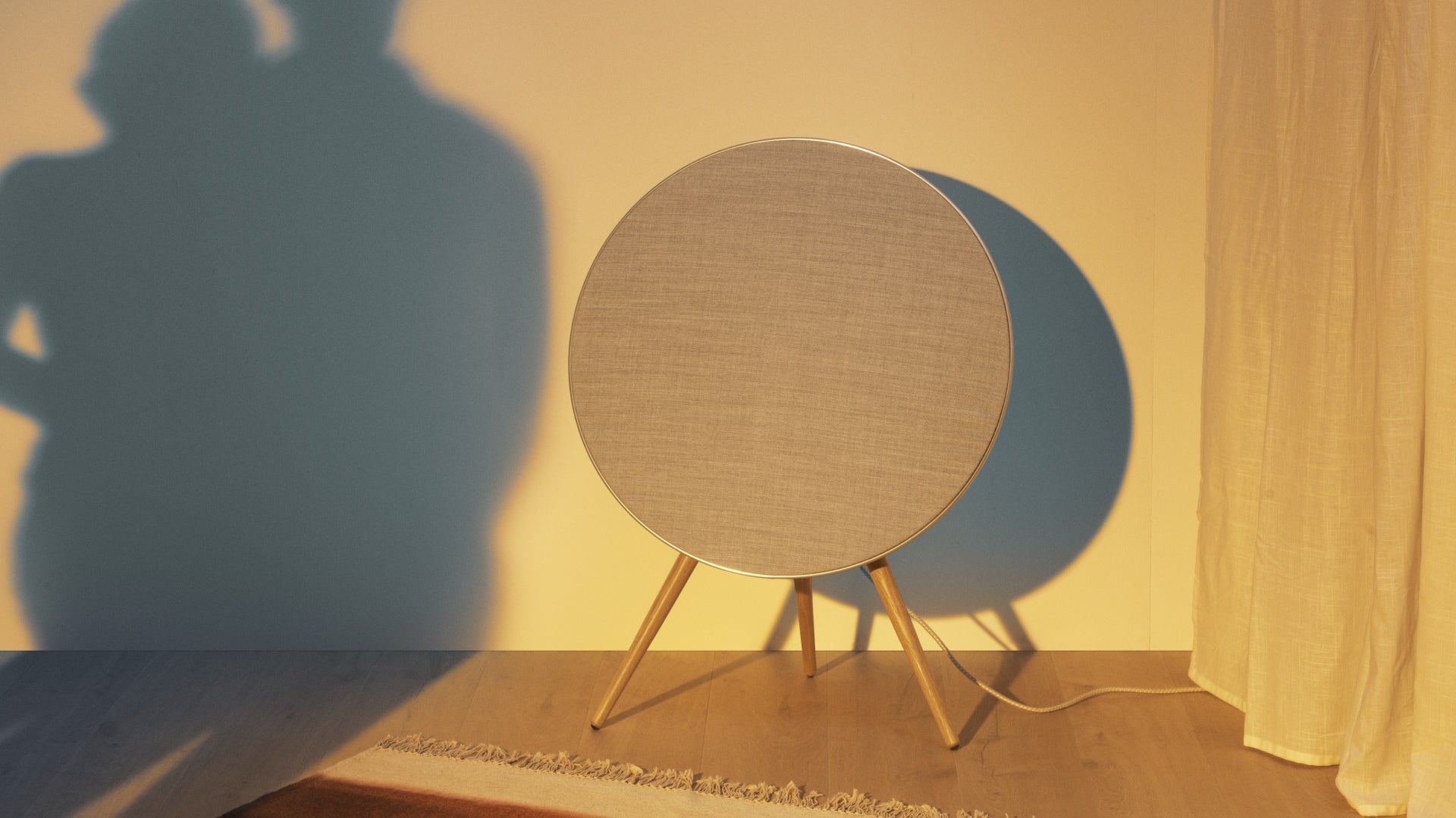 Stimmungsvolle Lifestyle-Aufnahme des Bang & Olufsen Beosound A9 Lautsprechers bei tiefstehender Abendsonne. Der ikonische, kreisrunde Lautsprecher mit heller Textilbespannung und Eichenholzbeinen wirft einen markanten Schatten an die Wand, während warmes, goldenes Licht die Szene auf dem hellen Holzfußboden flutet.