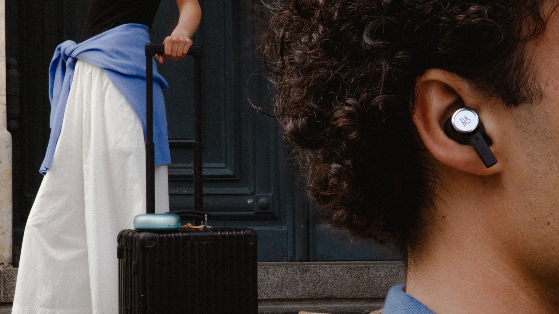 Lifestyle-Aufnahme der Bang & Olufsen Beoplay Eleven In-Ear-Kopfhörer in Black Anthracite während einer Reise. Ein Mann trägt den Earbud mit markantem Aluminium-Ring; im Hintergrund ist eine Frau mit einem schwarzen Aluminium-Koffer zu sehen, auf dem ein Beosound A1 in Eucalyptus Green liegt.