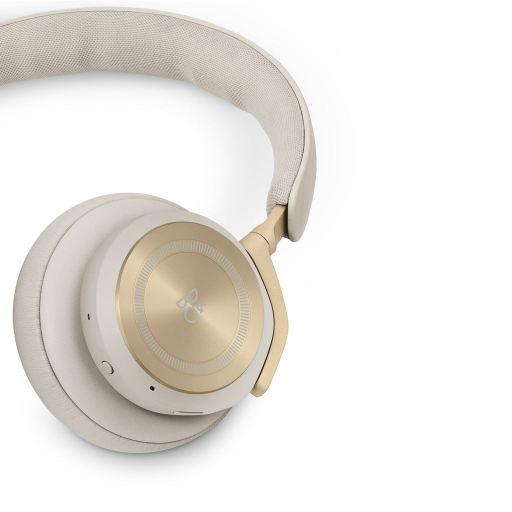 Detailaufnahme des Bang & Olufsen Beoplay HX in Gold Tone: Fokus auf die goldene Aluminium-Ohrmuschel mit B&O-Logo, das weiche Lammleder-Ohrpolster und den strukturierten Textil-Bezug an der Innenseite des Kopfbügels.