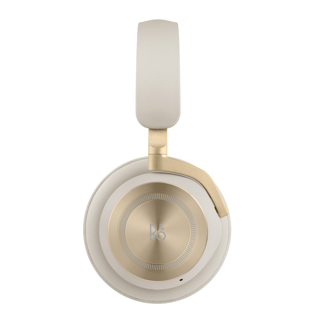 Frontale Seitenansicht des Bang & Olufsen Beoplay HX Over-Ear-Kopfhörers in der Farbe Sand. Der Fokus liegt auf der kreisrunden, goldfarbenen Aluminium-Ohrmuschel mit feiner Rillenstruktur und dem B&O-Logo, die an einem sandfarbenen Lammleder-Polster befestigt ist.